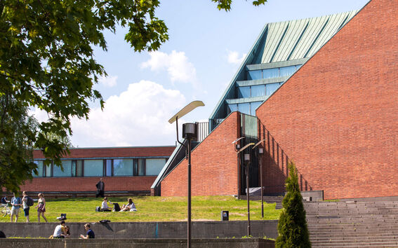People relax on a grassy area near a modern building with red brick walls and large glass windows under a blue sky.