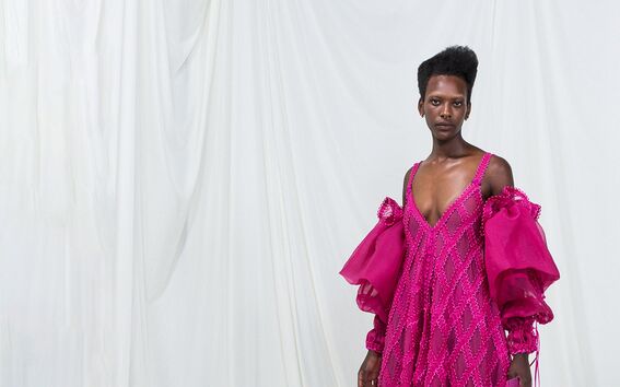Person wearing a bright pink dress with large puff sleeves, standing against a white draped background.