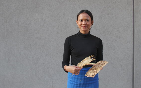 Henkilö mustassa paidassa ja sinisessä hameessa pitää kahta teksturoitua materiaalia käsissään, taustalla harmaa seinä.