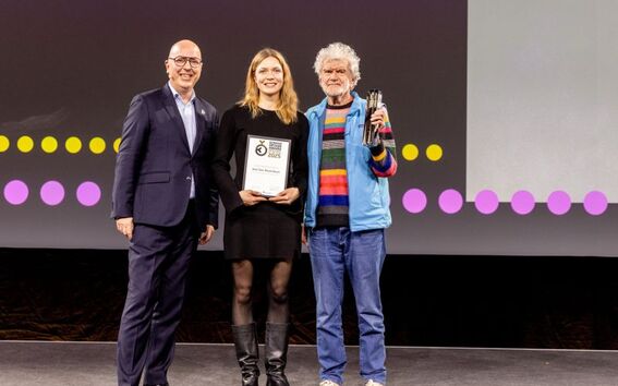 Three people standing on stage, one holding a plaque and another holding a trophy. Background has yellow and pink dots.