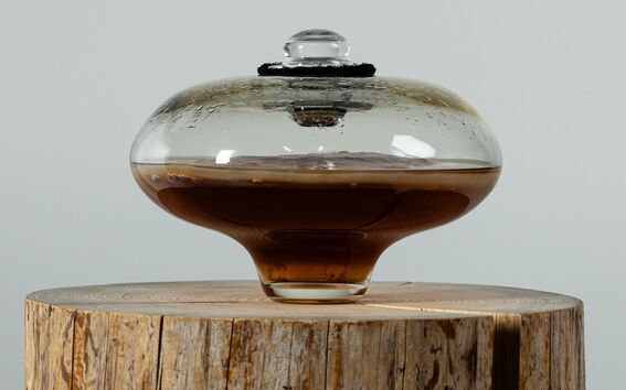 A glass container with a lid, filled with brown liquid, placed on a wooden stump against a grey background.