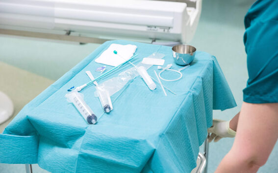 Lääketieteellisiä välineitä turkoosilla pöydällä, kuten ruiskuja, sideharsoa ja metallikulho. Henkilö turkooseissa vaatteissa lähellä.