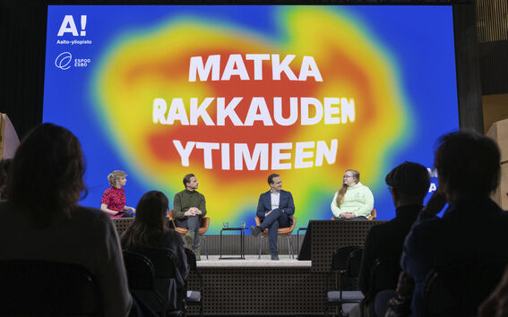 Neljä ihmistä istuu lavalla värikkään taustan edessä, jossa on suomenkielistä tekstiä. Yleisön jäsenet istuvat etualalla.