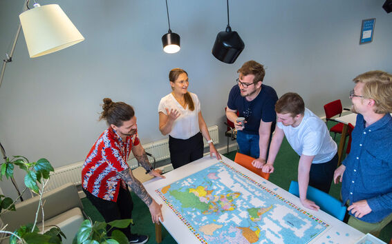 Viisi ihmistä seisoo pöydän ympärillä, jossa on maailman kartta -palapeli. Huoneessa on moderni valaistus ja vihreä matto.