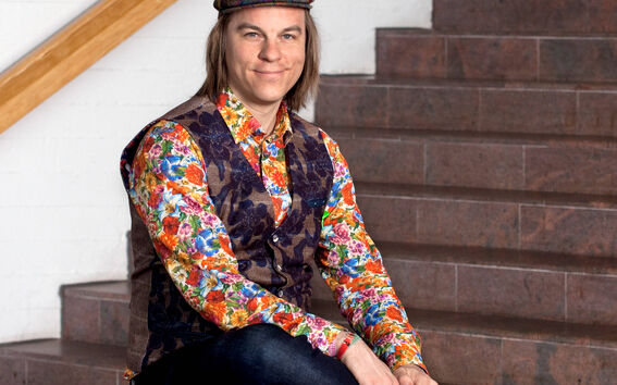 Henkilö istuu pyöreän pöydän päällä, yllään värikäs kukkapaita, tumma liivi, farkut ja ruudullinen hattu. Taustalla portaat.