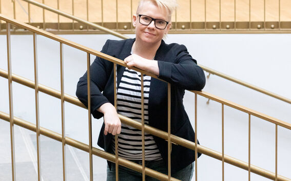 Henkilö nojaa kaiteeseen, yllään musta bleiseri, raidallinen paita ja farkut. Taustalla portaat ja puulattia.