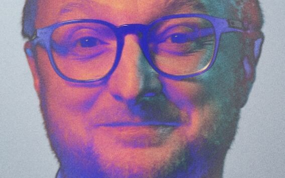 A person with short dark hair wearing a white shirt. The background is grey.