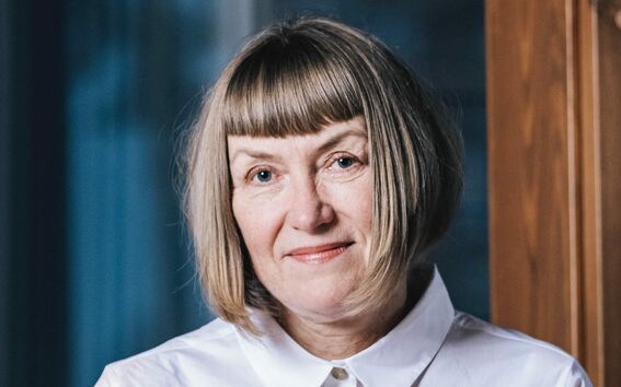 A person with short blonde hair wearing a white collared shirt, standing indoors near a wooden surface.