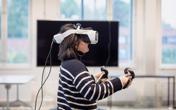 Person using a VR headset and controllers in a room with large windows and a black screen.