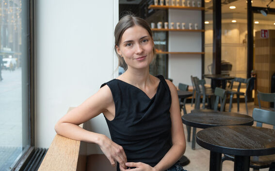 En person i svart ärmlös topp sitter på ett kafé nära ett fönster. Tomma bord och stolar syns i bakgrunden.