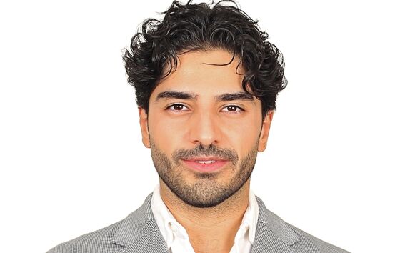 A person with curly hair wearing a grey blazer, white shirt, and blue pocket square, standing against a white background.