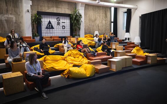 People sitting on tiered seating with yellow bean bags, laptops, and wooden tables in a modern room with plants.
