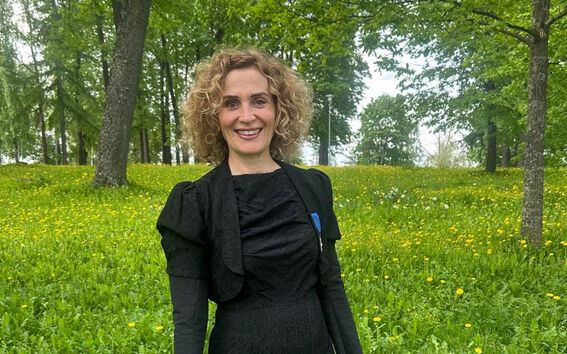 Eeva Furman in a black dress and gloves standing in a field with yellow flowers and trees in the background.