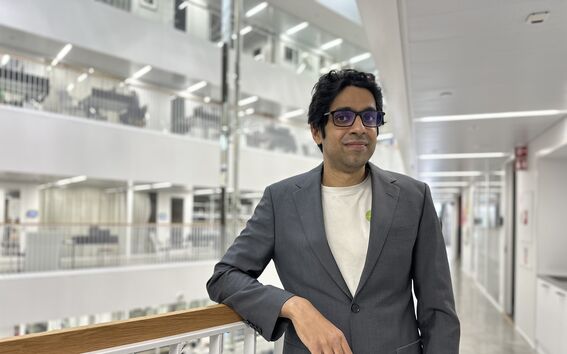 A person in a grey suit and white shirt stands in a modern, multi-storey building with glass and metal railings.