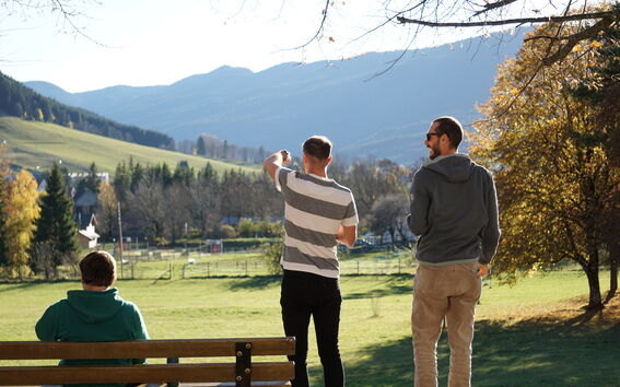 Kolme ihmistä puistossa, taustalla vuoria. Yksi istuu penkillä, kaksi seisoo maisemaa katsellen.