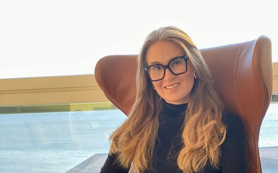 Pitkähiuksinen henkilö istuu ruskeassa tuolissa, yllään musta paita ja harmaa hame. Takana ikkuna, josta näkyy maisema.