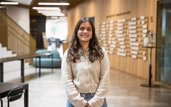 A person with long hair stands in a modern hallway with wooden walls and a staircase. They wear a beige cardigan and jeans.