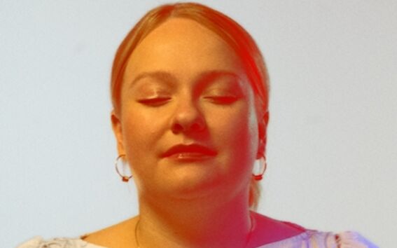 Enni Lähderinne with blonde hair wearing a light-coloured top with floral embroidery, standing against a plain background.