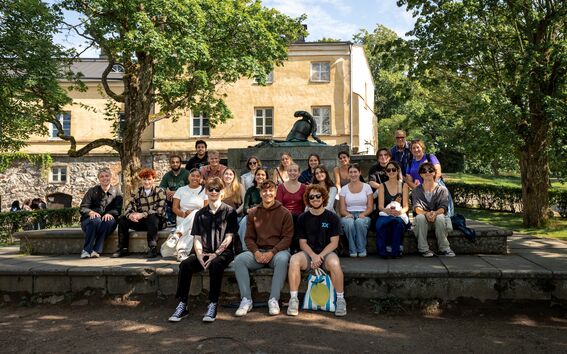 A group of people sitting on stone steps in a park with trees and a yellow building in the background.