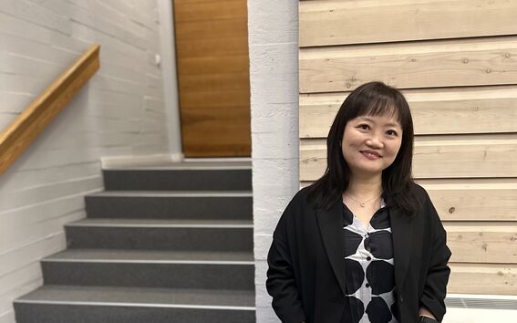 A person stands indoors near a staircase, wearing a black outfit with a patterned shirt. The walls are white and wooden.