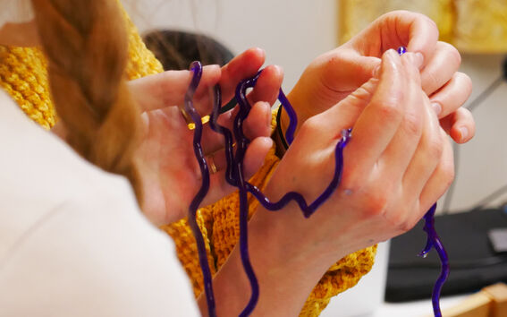A person with a braid holds purple alginate string. They wear a yellow knitted garment.