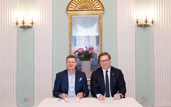 Kaksi miestä puvuissa istuu pöydän ääressä asiakirjojen kanssa. Takana on peili ja maljakko kukilla.