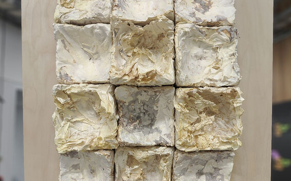 A wooden frame holds twelve crumpled, white paper-like squares arranged in a 3x4 grid. Background is blurred.