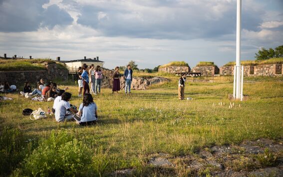 Ihmisiä on kokoontunut ulkona nurmikentälle, taustalla kivirakennelmia pilvisen taivaan alla.