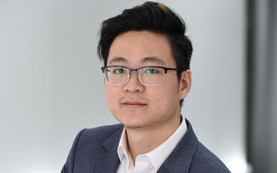 A person in a grey suit and white shirt stands with hands clasped in a modern, bright room.