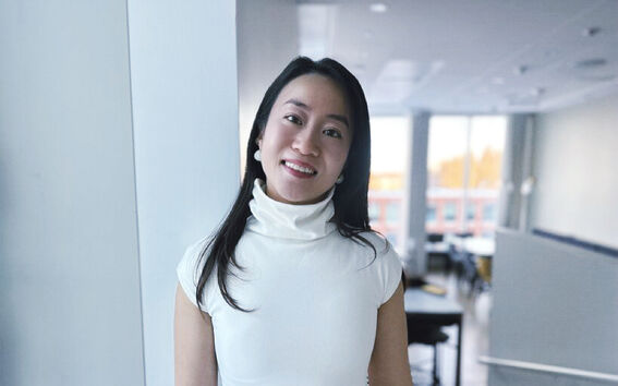 A person with long dark hair wearing a white turtleneck top stands indoors near a large window.