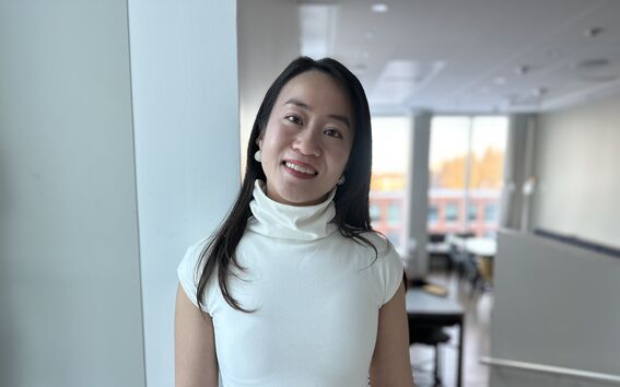 A person wearing a white turtleneck top and dark trousers stands indoors with a blurred background.
