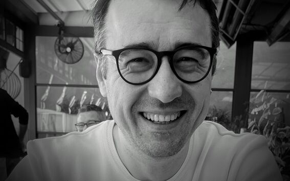 A person wearing a light-coloured shirt is sitting indoors near a window with plants outside.