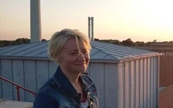 Person standing on a rooftop at sunset, wearing a denim jacket and striped shirt. Industrial buildings in the background.