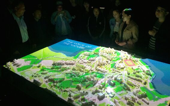 A group of people stand around a lit 3D model of a landscape with buildings and greenery. The model displays 'THE VOICE OF THE NATURE'.
