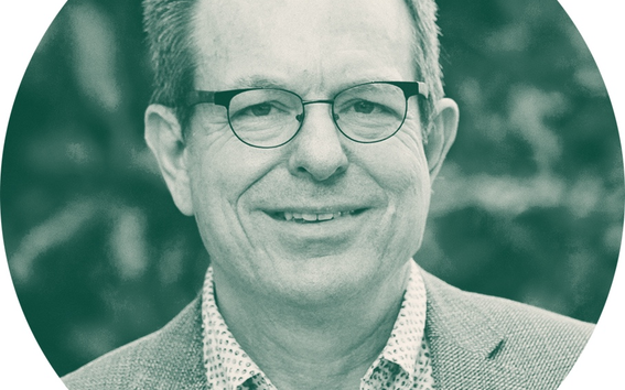 A person wearing a grey blazer and a white shirt with small dark patterns. The background is blurred greenery.