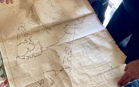 Several people examine an old map spread out on a floral tablecloth. One person points at a location on the map.