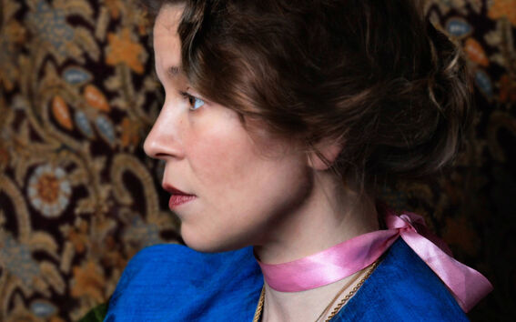 Henkilö, jolla on ruskeat hiukset sidottuna taakse, vaaleanpunainen nauha ja sininen vaate. Taustalla kuvioitu kangas.