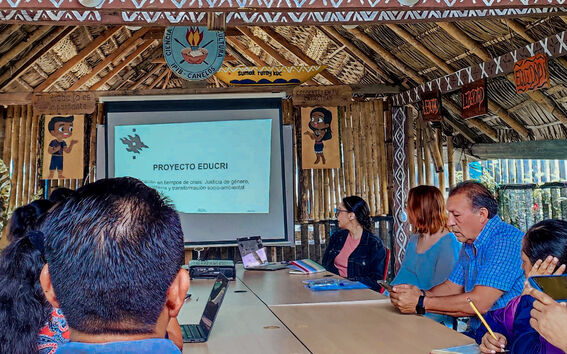 People in a bamboo hut attending a presentation titled 'Proyecto Educri'. Various posters and decorations are visible.