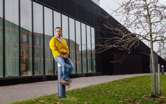 Person in a yellow jacket and jeans sits on a bollard outside a modern building with large windows and a tree nearby.