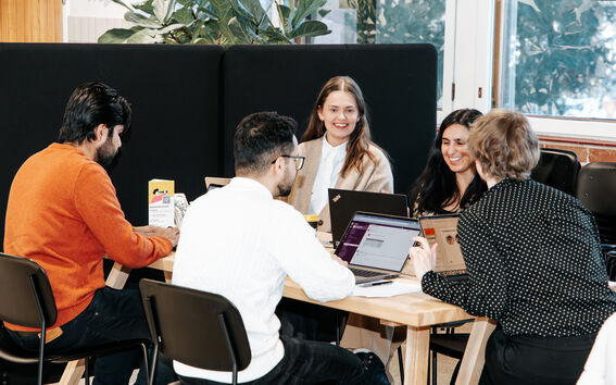 Five people sitting at a table with laptops, engaged in discussion. Plants and large windows are in the background.