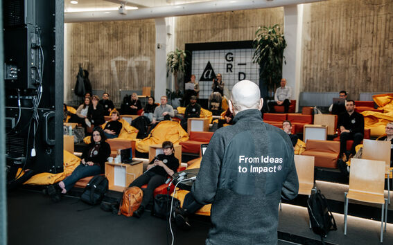 A person presenting to an audience in a modern lecture hall with yellow bean bags and wooden chairs.