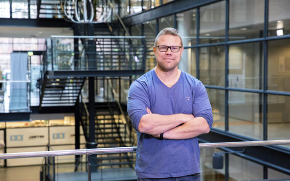 Henkilö sinisessä paidassa seisoo kädet ristissä modernissa rakennuksessa, jossa on lasiseinät ja metalliset portaat.