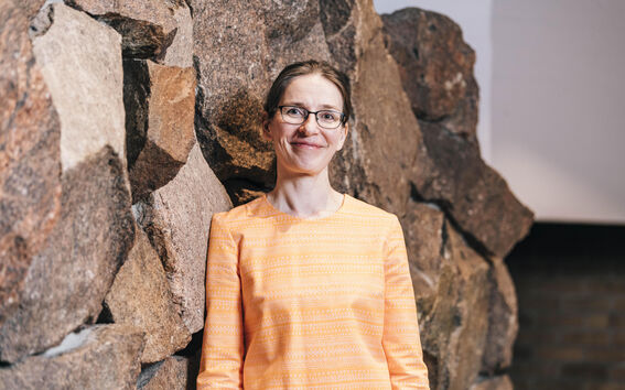 Henkilö oranssissa kuvioidussa paidassa seisoo suuren kiviseinän edessä.