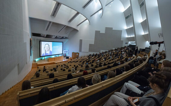 Suuri luentosali kaarevilla istuimilla, esitysnäyttö ja yleisö seuraamassa puhujaa.