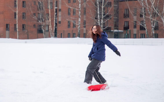Person in a blue coat pulling a red sled in a snowy area with brick buildings and bare trees in the background.