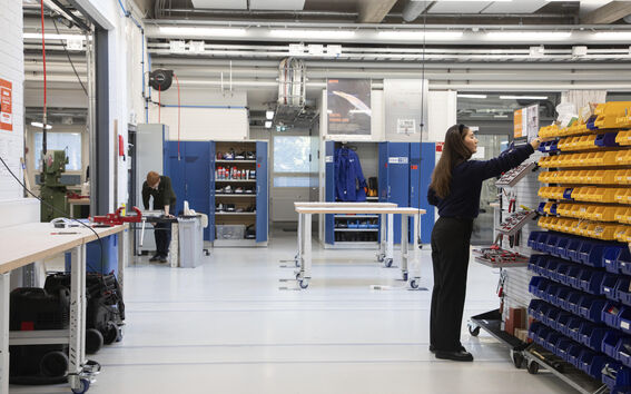 Työpaja, jossa kaksi ihmistä työskentelee. Toinen on pöydän ääressä, toinen järjestelee tavaroita keltaisissa ja sinisissä laatikoissa.