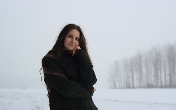Pitkähiuksinen henkilö tummassa neuleessa seisoo lumisella pellolla, taustalla paljaita puita.