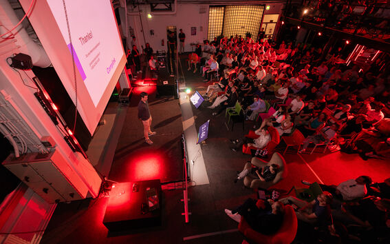 A speaker presents to a large audience in a dimly lit room with red lighting.