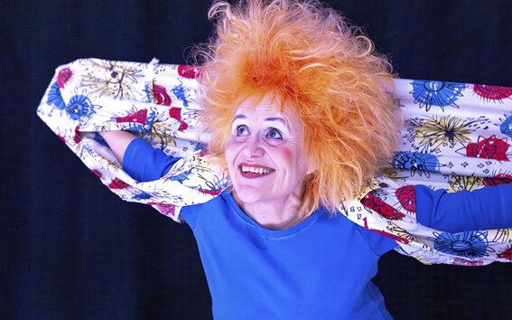 Henkilö, jolla on kirkkaan oranssit hiukset, sininen paita ja värikäs kuvioitu kangas tummalla taustalla.