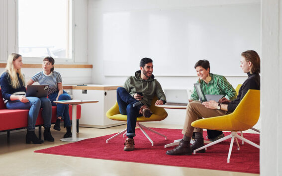 Five people are sitting in a modern office space with laptops and notebooks, discussing. There is a red carpet and colourful furniture.